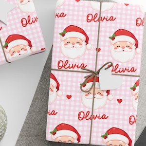 May include: Gift-wrapped presents with pink Santa Claus patterned paper, tied with twine and adorned with white tags. The wrapping paper features a repeating pattern of Santa faces, hearts, and the name "Olivia" on a checkered background. A ball of twine and scissors are also visible.