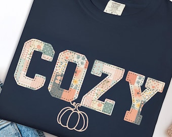 May include: Navy blue t-shirt with the word "COZY" in a patchwork design with floral patterns and a pumpkin graphic. The shirt is folded next to a pair of blue jeans, white slip-on shoes, and a beige blanket.