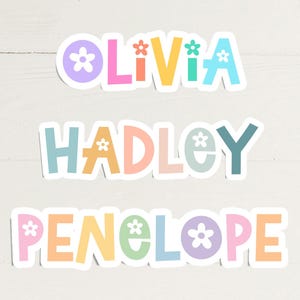 May include: Three colorful stickers with the names Olivia, Hadley, and Penelope. Each name is in a different pastel color, with a flower graphic replacing the letter "o". The stickers are on a white wooden surface.
