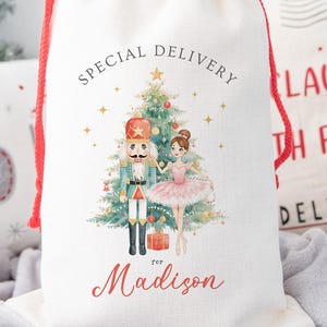 May include: A white drawstring gift bag with a red drawstring closure. The bag features a Christmas tree, a nutcracker, and a ballerina illustration. The text "Special Delivery" and "for Madison" are printed on the bag.