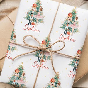 May include: Wrapped gift with white paper featuring a repeating pattern of a Christmas tree, a nutcracker, and a ballerina. The name "Sophia" is printed in red. The gift is tied with twine and sits on a brown paper package.