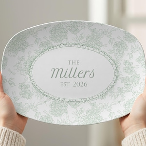 May include: An oval serving platter with a floral design in light green and white. The center features an oval with the text "The Millers EST. 2026" in a script font. The platter is held up by two hands.