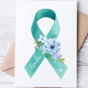May include: A greeting card featuring a teal ribbon adorned with blue and purple flowers. The word "Linda" is written on the ribbon. The card is on a white background, with a brown envelope behind it.
