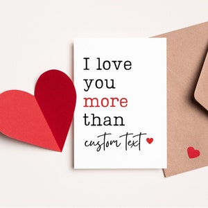 May include: A white card with the text "I love you more than custom text" and a small red heart. The card is next to a brown envelope and a red paper heart.