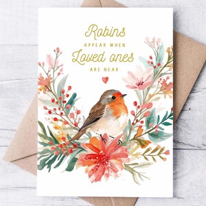 May include: Greeting card featuring a watercolor illustration of a robin perched amidst a floral wreath. The card reads "Robins appear when loved ones are near" in gold script. The card is set against a brown envelope.