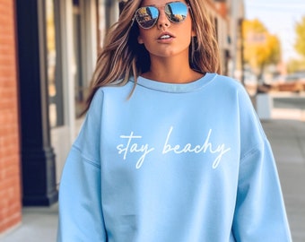 May include: A light blue sweatshirt with the text "stay beachy" in white lettering. The model is wearing white shorts and sunglasses.