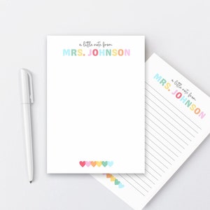 May include: White notepad with the text "a little note from MRS. JOHNSON" in pastel colors. A white pen is next to the notepad. The bottom of the notepad has a row of colorful hearts.
