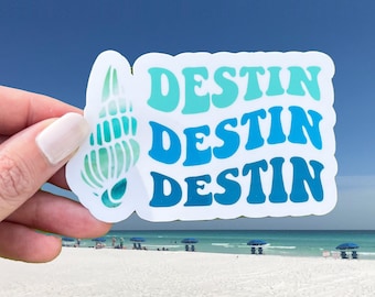 May include: A white sticker with the word "Destin" repeated three times in blue and green lettering. The sticker is held in a hand against a background of a beach with blue water and white sand.