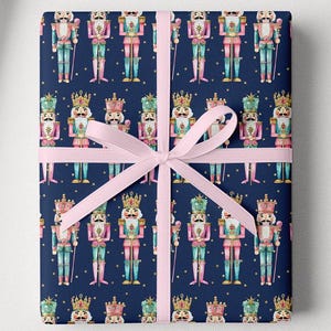 May include: Gift wrap featuring a repeating pattern of colorful nutcracker soldiers on a navy blue background. A wrapped gift with a pink bow is in the center. A roll of matching wrapping paper is on the left.