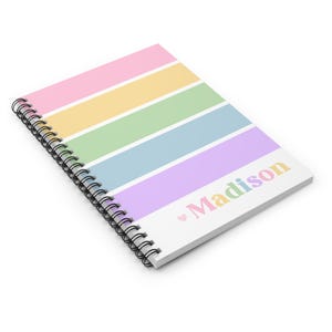 Op de afbeelding: Een spiraalgebonden notitieboek met een pastelkleurige regenboog gestreepte omslag. Het notitieboek heeft een wit gedeelte met de naam "Madison" in pastelkleuren en een klein roze hart.