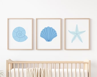 Puede incluir: Tres impresiones enmarcadas con ilustraciones en azul claro con temática marina. Las impresiones presentan una concha en espiral, una concha en forma de abanico y una estrella de mar. La obra de arte se exhibe sobre una cuna de madera.