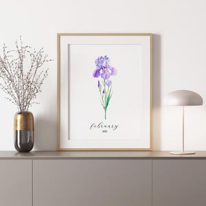 May include: A framed print featuring a watercolor illustration of a purple iris flower. The word "February" is written in cursive below the flower, with "IRIS" below that. The print is in a gold frame and is displayed on a gray cabinet.