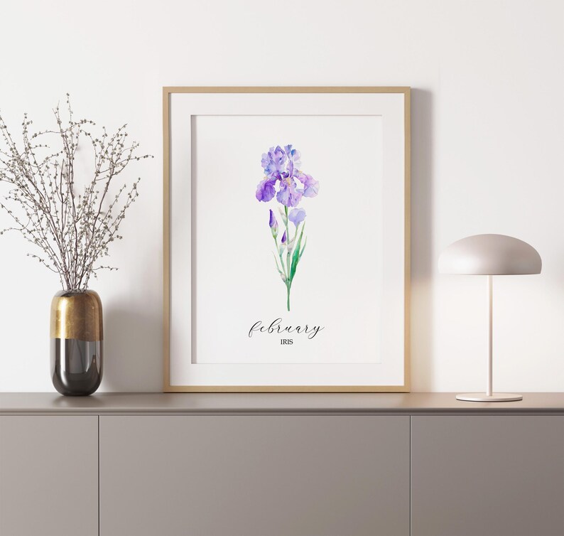 May include: A framed print featuring a watercolor illustration of a purple iris flower. The word "February" is written in cursive below the flower, with "IRIS" below that. The print is in a gold frame and is displayed on a gray cabinet.