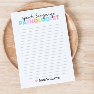 Peut inclure: Un bloc-notes blanc avec des lignes noires et le texte "speech language PATHOLOGIST" en couleurs arc-en-ciel. Le bas du bloc-notes a un c&oelig;ur rose et le texte "Miss Williams".