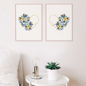 May include: Two framed art prints featuring floral heart designs. Each print has a gold heart outline filled with blue and yellow watercolor flowers. One print says "love" and the other says "faith" in gold script. The frames are a light pink color.