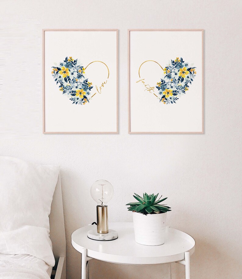 May include: Two framed art prints featuring floral heart designs. Each print has a gold heart outline filled with blue and yellow watercolor flowers. One print says "love" and the other says "faith" in gold script. The frames are a light pink color.