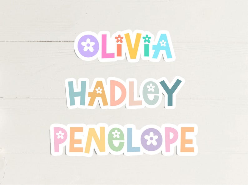 May include: Three colorful stickers with the names Olivia, Hadley, and Penelope. Each name is in a different pastel color, with a flower graphic replacing the letter "o". The stickers are on a white wooden surface.