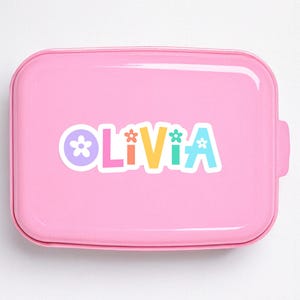 May include: A pink lunchbox with rounded edges. The word "Olivia" is printed on the front in colorful letters, each with a flower accent. The lunchbox has a handle on the side.