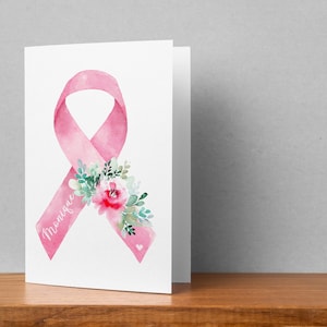 May include: A folded white card featuring a watercolor pink ribbon with a floral arrangement and the name "Monique" in script. The ribbon is a symbol of breast cancer awareness. The card is on a wooden surface.