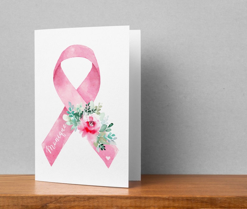 May include: A folded white card featuring a watercolor pink ribbon with a floral arrangement and the name "Monique" in script. The ribbon is a symbol of breast cancer awareness. The card is on a wooden surface.