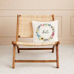 May include: A white pillow with a floral wreath design and the name "Kate" in black script. The pillow is on a light brown wicker chair.