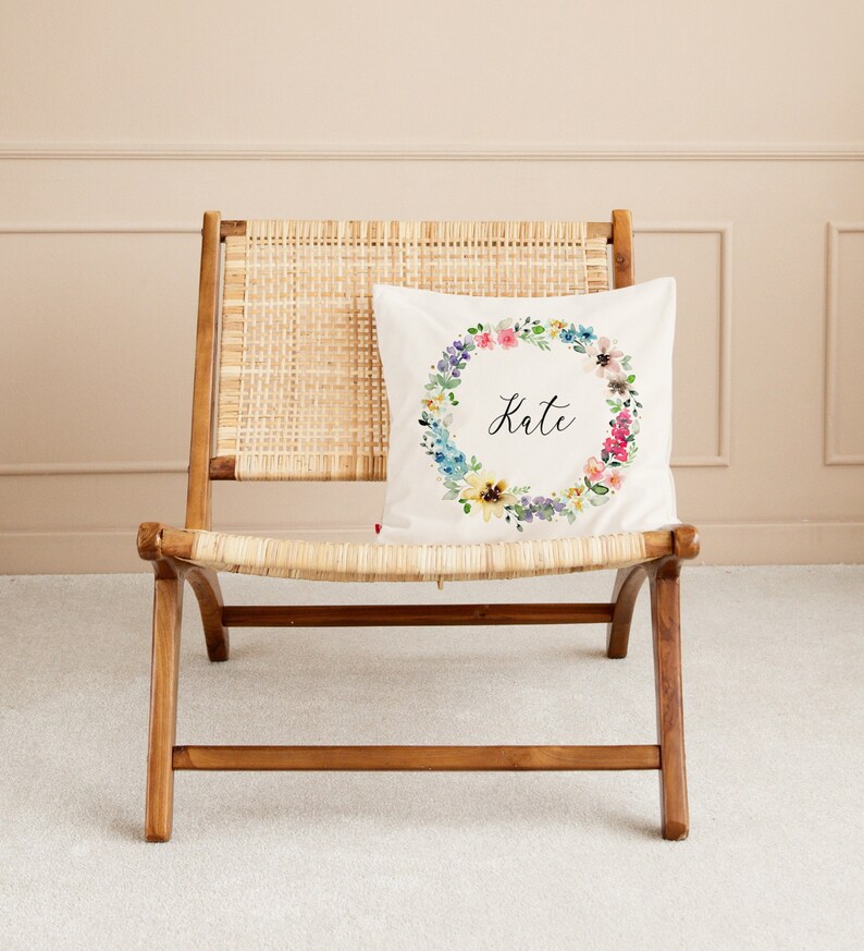 May include: A white pillow with a floral wreath design and the name "Kate" in black script. The pillow is on a light brown wicker chair.
