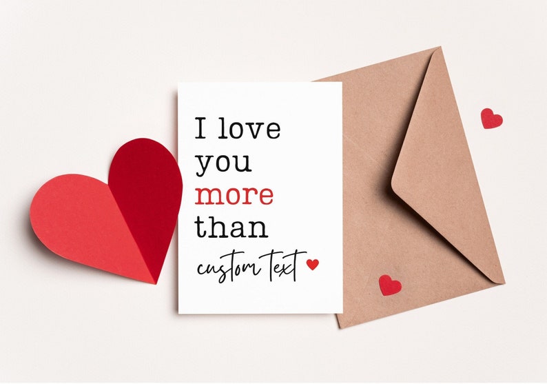 May include: A white card with the text "I love you more than custom text" and a small red heart. The card is next to a brown envelope and a red paper heart.