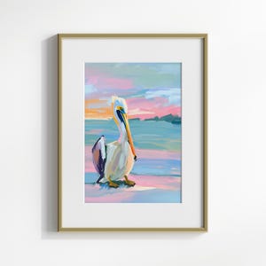 May include: A framed painting of a pelican standing on a beach at sunset. The pelican is white with a long orange beak and blue and purple accents. The background is a soft pink and blue.