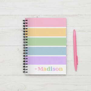 Op de afbeelding: Een spiraalgebonden notitieboek met een pastelkleurig regenboogstreepontwerp en de naam "Madison" in regenboogkleuren. Een roze pen ligt naast het notitieboek op een wit houten oppervlak.
