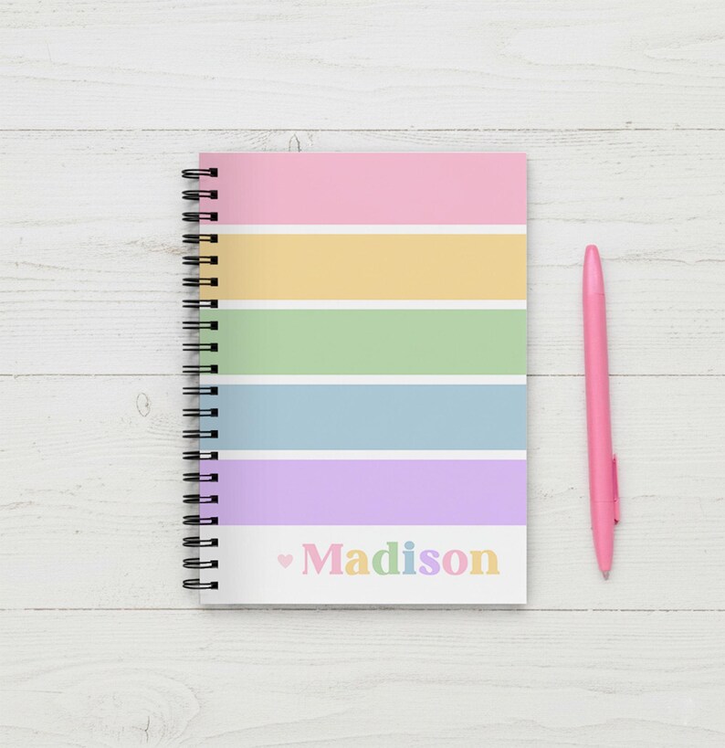 Op de afbeelding: Een spiraalgebonden notitieboek met een pastelkleurig regenboogstreepontwerp en de naam "Madison" in regenboogkleuren. Een roze pen ligt naast het notitieboek op een wit houten oppervlak.