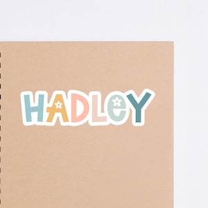 May include: A colorful sticker with the name "HADLEY" in a playful font. The letters are in shades of blue, orange, and pink, with a small flower graphic. The sticker is placed on a spiral-bound notebook with a tan cover.