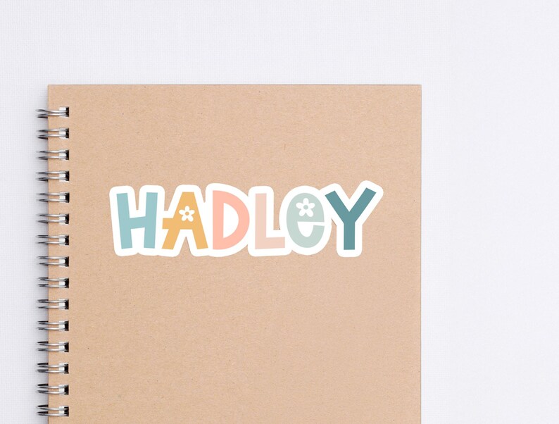 May include: A colorful sticker with the name "HADLEY" in a playful font. The letters are in shades of blue, orange, and pink, with a small flower graphic. The sticker is placed on a spiral-bound notebook with a tan cover.