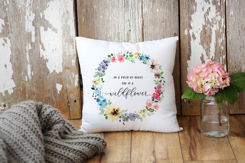 May include: White throw pillow with a watercolor floral wreath and the text "In a field of roses she is a wildflower".