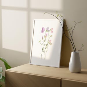 May include: A framed watercolor print featuring delicate purple and pink sweet pea flowers. The white frame leans against a wall, next to a white vase holding branches. The artwork is displayed on a wooden surface.