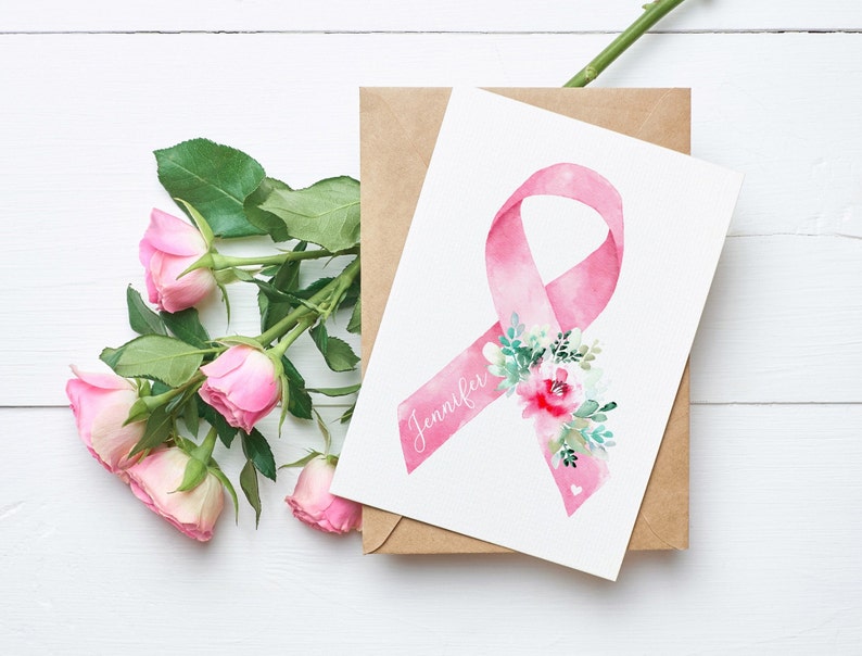 May include: A watercolor greeting card featuring a pink ribbon with the name "Jennifer" and floral accents. The card is on a brown envelope, with pink roses and green leaves in the background.