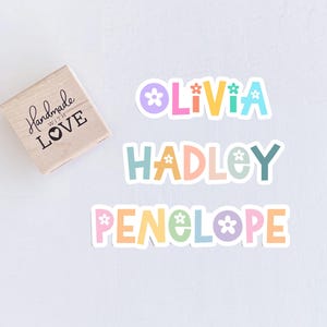May include: A wooden stamp with the text "Handmade with Love" and three colorful name stickers: "Olivia", "Hadley", and "Penelope". Each name is in a different pastel color with flower accents.