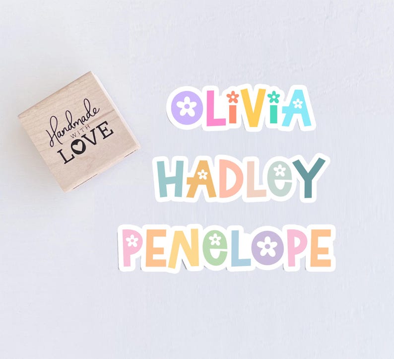 May include: A wooden stamp with the text "Handmade with Love" and three colorful name stickers: "Olivia", "Hadley", and "Penelope". Each name is in a different pastel color with flower accents.