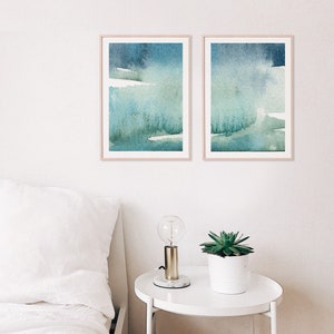 May include: Two framed abstract watercolour paintings with shades of blue and green. The paintings are hung on a white wall above a white bedside table with a lamp and a potted plant.