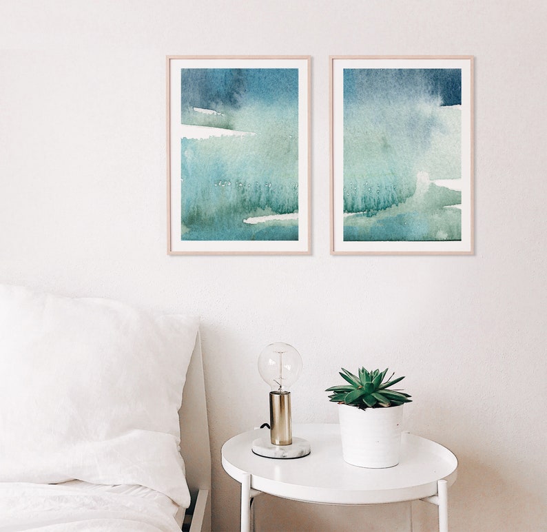 May include: Two framed abstract watercolour paintings with shades of blue and green. The paintings are hung on a white wall above a white bedside table with a lamp and a potted plant.
