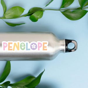 May include: A silver water bottle with a black cap and the name "PENELOPE" in pastel colors. The word is decorated with flower and star graphics. Green leaves and a light blue background complete the image.