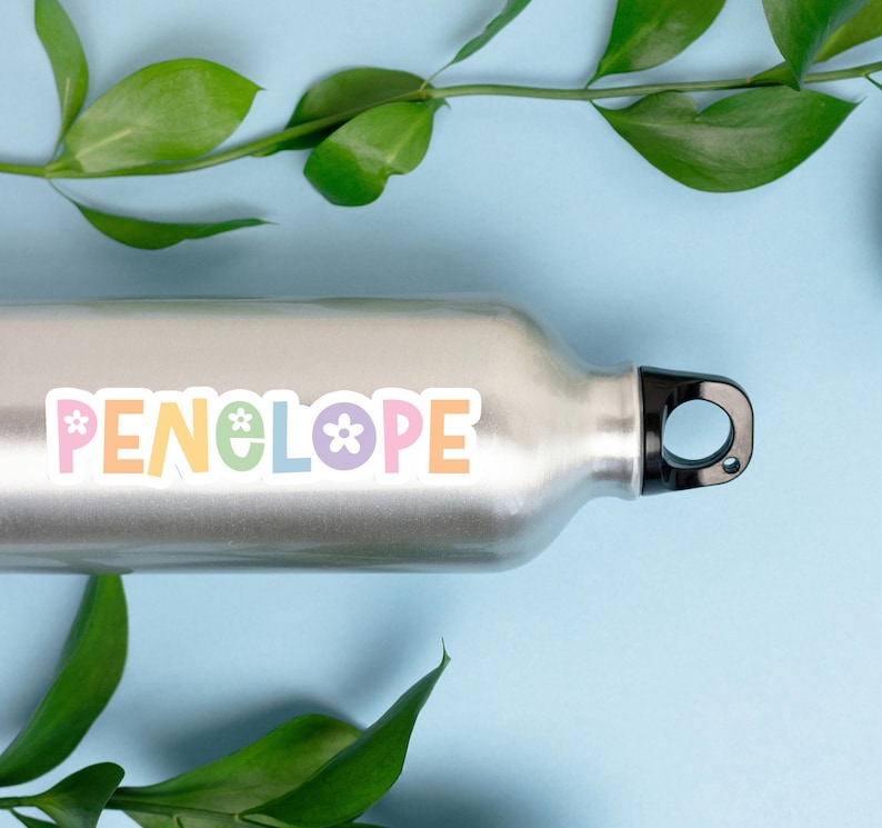 May include: A silver water bottle with a black cap and the name "PENELOPE" in pastel colors. The word is decorated with flower and star graphics. Green leaves and a light blue background complete the image.