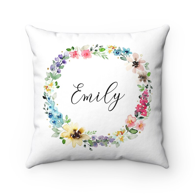 May include: White pillow with a floral wreath design in shades of pink, blue, yellow, and green. The wreath surrounds the name "Emily" in black cursive font.