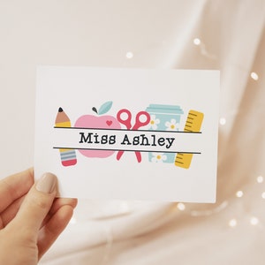 May include: A white card with a pink apple, a yellow pencil, a pair of pink scissors, a blue cup with white flowers, and a yellow ruler. The text "Miss Ashley" is printed on the card.