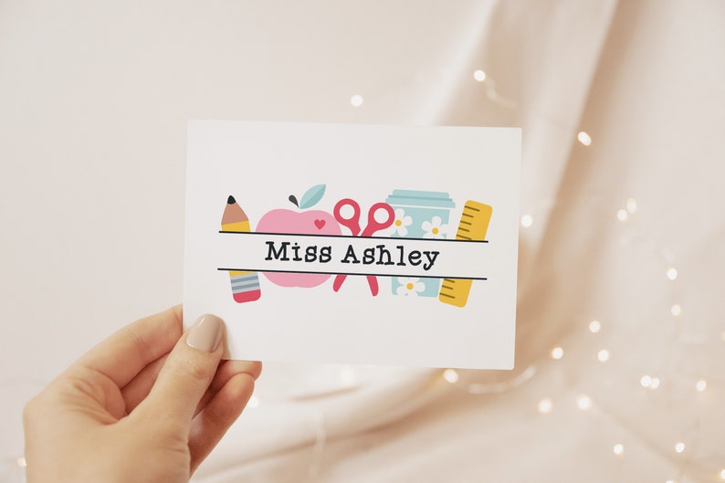 May include: A white card with a pink apple, a yellow pencil, a pair of pink scissors, a blue cup with white flowers, and a yellow ruler. The text "Miss Ashley" is printed on the card.