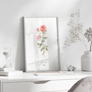 May include: A framed watercolor print featuring pink roses, with the text "june ROSE". The artwork is displayed on a white desk, alongside a vase of white flowers, an hourglass, and a small notebook. The scene is bright and airy.