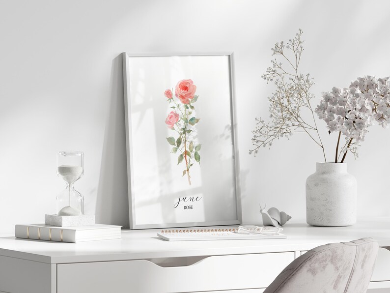May include: A framed watercolor print featuring pink roses, with the text "june ROSE". The artwork is displayed on a white desk, alongside a vase of white flowers, an hourglass, and a small notebook. The scene is bright and airy.