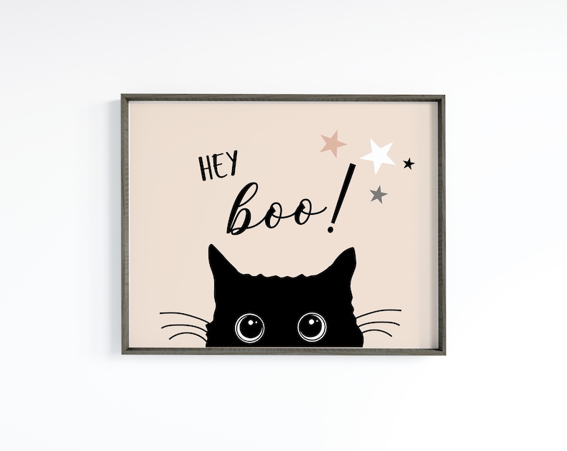 May include: A black cat with big eyes peeks over the top of a framed print with a peach background. The print says "Hey Boo!" with stars above the text.