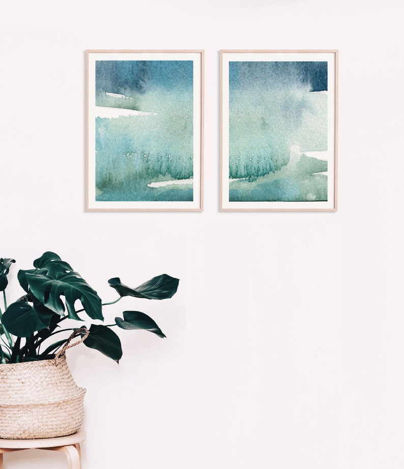 May include: Two framed abstract watercolour paintings in shades of blue and green. The paintings are hung on a white wall.