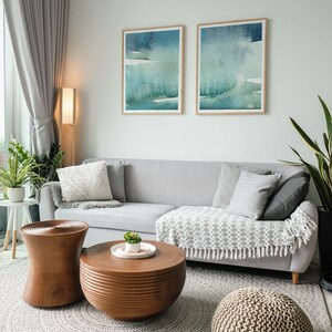 May include: A living room with a grey sectional sofa, two framed abstract watercolour paintings, a wooden coffee table, a wooden side table, a knitted pouffe, and a grey rug.
