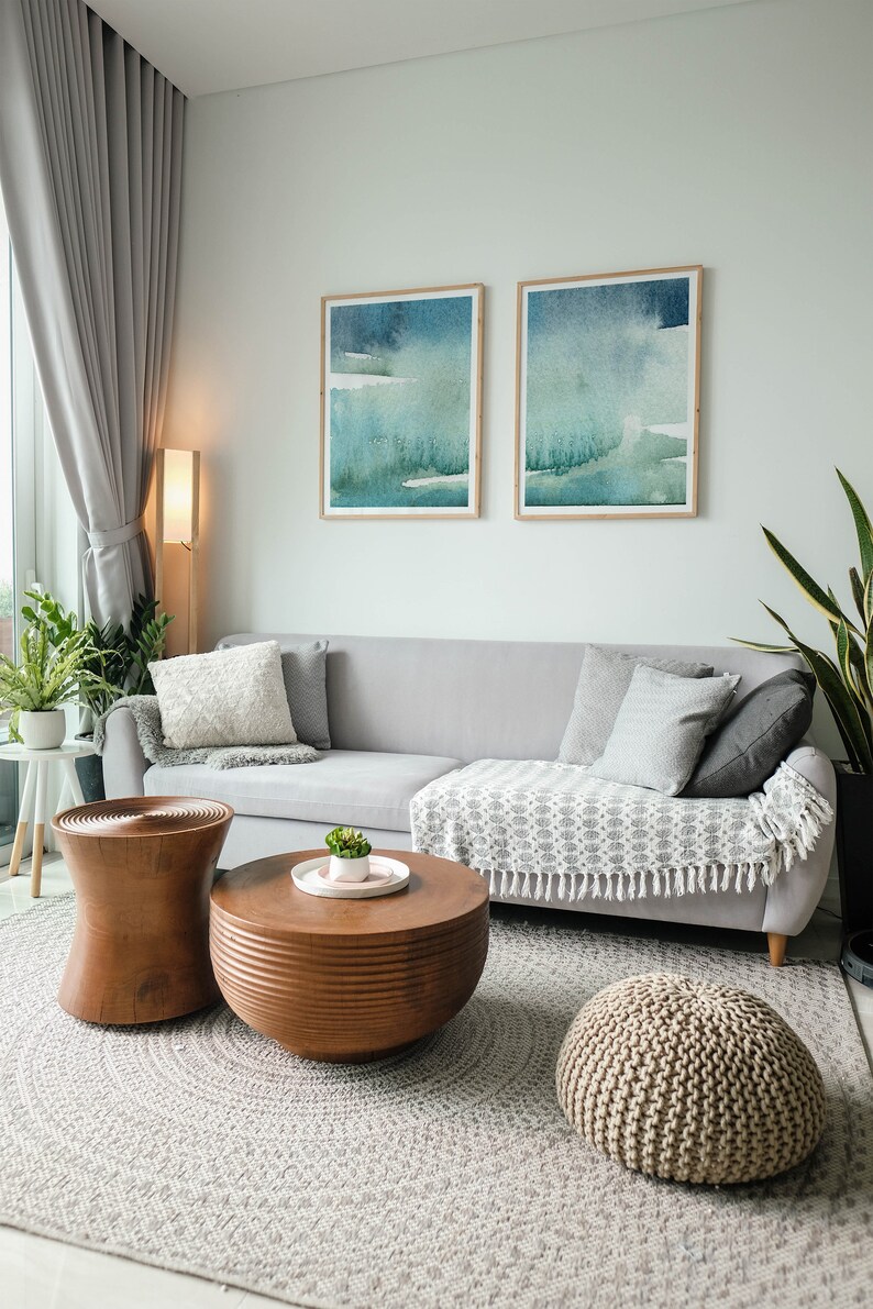 May include: A living room with a grey sectional sofa, two framed abstract watercolour paintings, a wooden coffee table, a wooden side table, a knitted pouffe, and a grey rug.